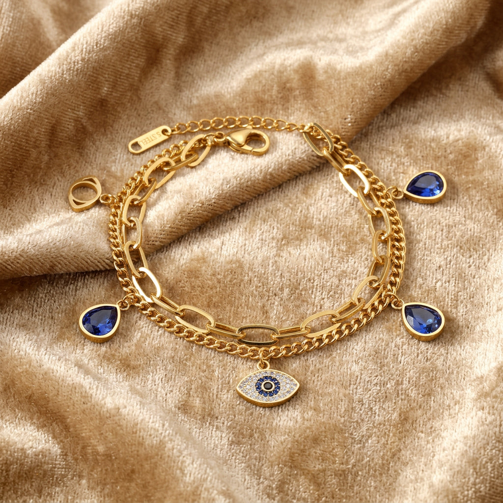 Protected in Light gold-tone stainless steel charm bracelet with layered chains, blue cubic zirconia teardrops and an evil eye charm on champagne fabric.